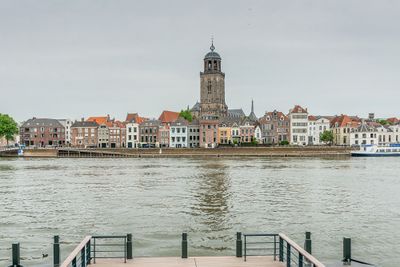 Stadsaanzicht van Deventer. In de voorgrond water en in de achter grond een huizenrij met een kerk in het midden.