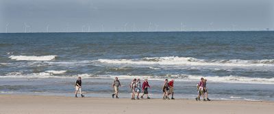Strand bij Bergen. Op het strand loopt een groep mensen. Ver in de zee staan windmolens.