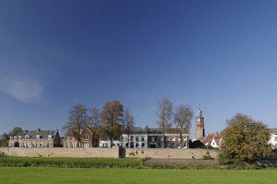 Stadaanzicht van Buren. Voor de huizenrij staat een stadsmuur met een grasveld ervoor. Rechts is een kerkje te zien.