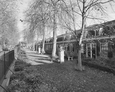 Zwart wit foto van het Hofje Eleëmosynae van Oudmunster. Links een hekwerk en rechts een huizenrij. Daartussen een ruimte met struiken, bomen en pompen.