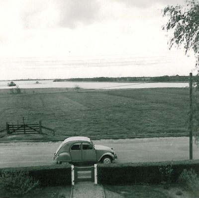 Oude zwart-wit foto van Haskerveen. In de voorgrond een weg waaroverheen een auto rijdt. Op de achtergrond is weiland te zien.