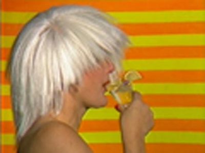 Still uit een video met daarop een vrouw met een grijze pruik die een coacktail drinkt tegen geel-oranje gestreepte achtergrond.