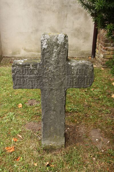 Een stenen grafkruis staat op een groen veld met op de achtergrond een muur.