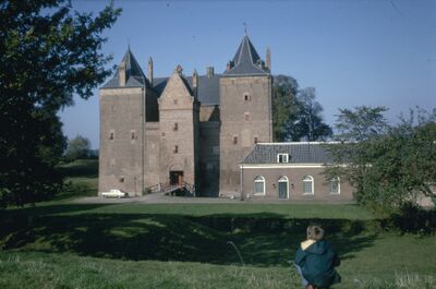 Twee gebouwen van het kasteel in het midden van de foto. Het grootste gebouw is voorzien van rechthoekige torens en een zwart dak met leien. Op de voorgrond een grasveldje.