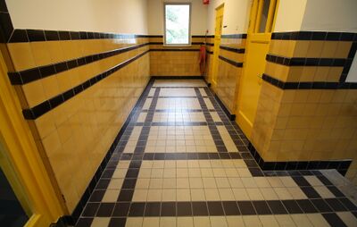 Gang in een Haags schoolgebouw. De vloer is met zwarte en witte tegels bedekt en de muur met gele en zwarte tegels.