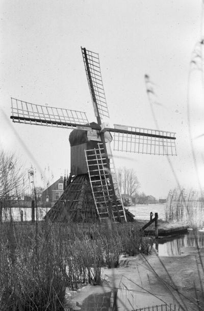 Zwartwit foto van de Haensmole op de oorspronkelijke locatie aan het Prinses Margrietkanaal/Pikmeer. Op de voorgrond water en begroeiing. Achter de molen water en in de verte bebouwing.