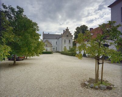 Foto van het binnenhof van het kasteel met rechts bijgebouwen. Rechtsvoor staat een jong boompje, links staat een grijze auto geparkeerd onder grotere bomen.