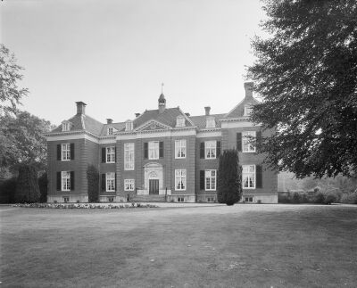 Zwart-witfoto van de voorgevel van het huis met een gazon in de voorgrond. In de omgeving staan bomen.