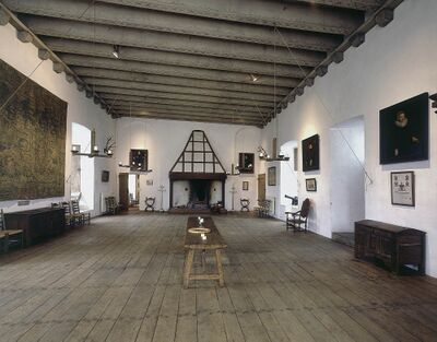 Een middeleeuws ogende kamer in een kasteel. De vloer is van hout en de kamer heeft witte, hoge muren.