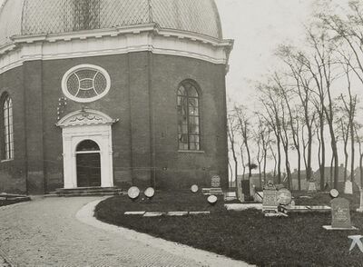 Kerkgebouw met veld vol grafstenen met daartussen trommels waarin kransen zitten