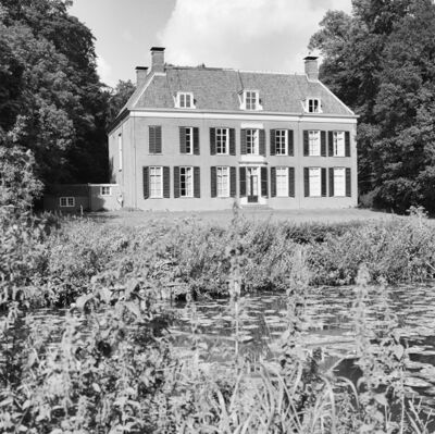 Zwart-witfoto van het huis met links een laag bijgebouw. In de voorgrond ligt een deels dichtgegroeide vijver en gazon daarachter. Het huis is omringd door bomen.