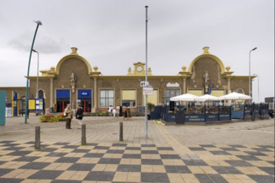 Foto van een bruin treinstation met rechts een terras afgescheiden door windschermen. In de voorgrond liggen tegels met een schaakbordpatroon, met houten paaltjes en een lantaarnpaal. Op het plein voor het station staan planten en lopen meerdere mensen rond.