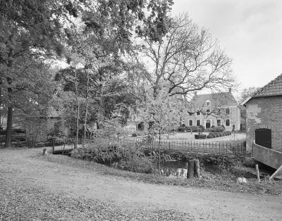 Zwart-witfoto met het kasteel en de tuin in de achtergrond met daarvoor de gracht met brug, deels zichtbaar achter jonge struiken en bomen. Aan weerszijden van de tuin staan bijgebouwen.