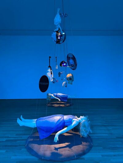 Mobiles met diverse voorwerpen en liggende vrouwfiguren in een blauw-aangelichte tentoonstellingsruimte.