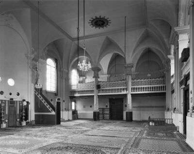Zwart-wit foto van de binnenkant van de moskee. De grond is bedekt met kleden en aan het hoge plafond hangen kroonluchters. Achterin is een vide.