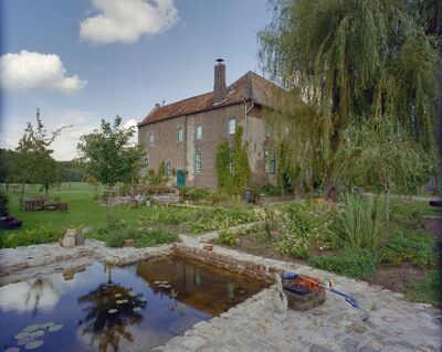 Huis Caldenbroek in het midden van de foto. Eromheen een tuin die vol in bloei staat. Rechts vooral gras met bomen. Vooraan in de foto ligt een vijver en een stuk aarde met struiken die roze, witte en oranje bloemen bevatten. Door de tuin loopt een stenen pad.