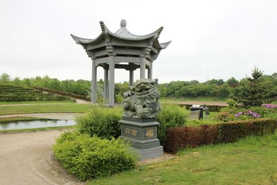Centraal een in grijze natuursteen uitgevoerd paviljoen en daarvoor een beeld van een leeuw alles in Chinese stijl.