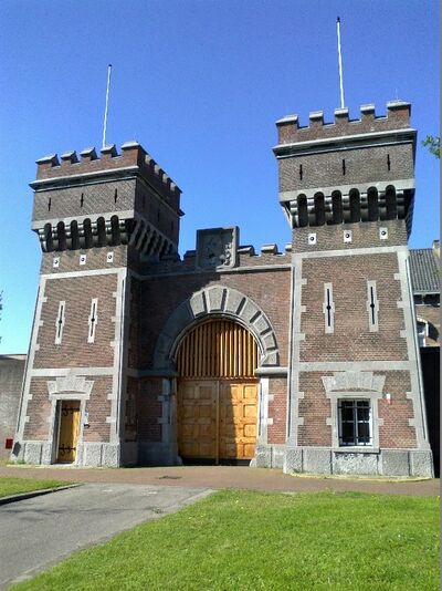 Toegangspoort naar de strafgevangenis in Scheveningen. Aan weerszijden van de poort staan twee grote bakstenen torens. De poortdeuren zijn van lichtbruin gelakt hout.
