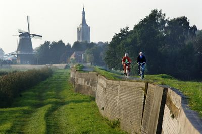 Stenendijk bij Hasselt. Rechts de met bakstenen verstevigde dijk waar twee fietsers fietsen. In de achtergrond een kerktoren en molen.