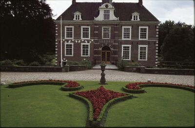 Foto van de siertuin voor de ingang van het kasteel, met lage heggen in geometrische vormen en rode planten. Tussen het kasteel en de tuin ligt een grindpad. Er loopt een brug van het pad naar de ingang van het kasteel.