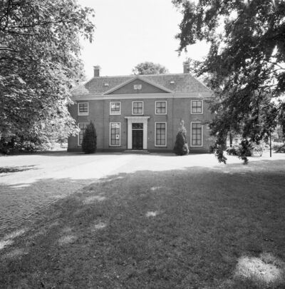 Zwart-witfoto van de ingangszijde van het landhuis. Het is omringd door gras en in de voorgrond van de foto bungelen aan weerszijden bloeiende boomtakken.