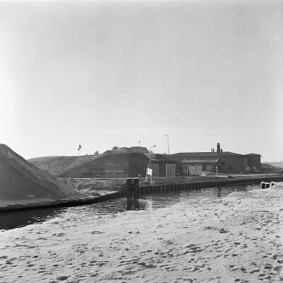 Zwart-wit foto van het fort met in de voorgrond water. Ernaast staat een nieuwer aangelegd gebouw met hekken ervoor.