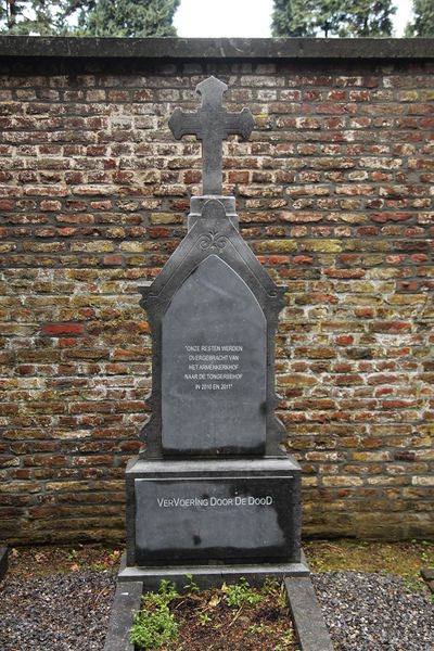 Een oud grafmonument met een nieuwe tekst staat voor een muur op een begraafplaats.