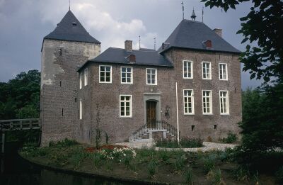 Foto van het kasteel en voorplein met beplante border en vlaggenstok. Links is de gracht en brug zichtbaar, de gracht is omringd door bomen.
