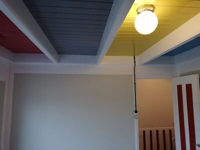 De dragende balken van het plafond zijn wit geschilderd. De stroken met planken tussen deze balken zijn uniform geschilderd in rood, lichtblauw en geel. Aan de rechterkant is een open deur zichtbaar, deze is wit geschilderd met rode verticale stroken.